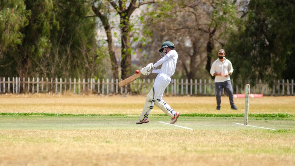 Batsman in white gear hits cricket ball on sunny outdoor field.