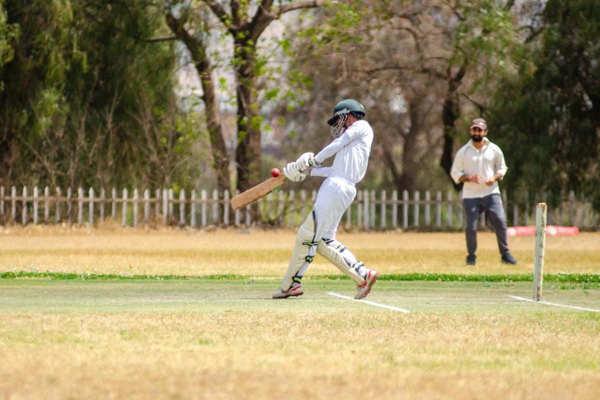 Batsman in white gear hits cricket ball on sunny outdoor field.